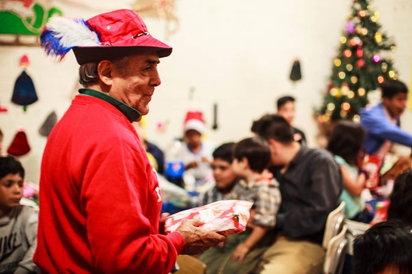 Mann in rotem Pullover mit Geschenk, umgeben von feiernden Kindern und einem Weihnachtsbaum.