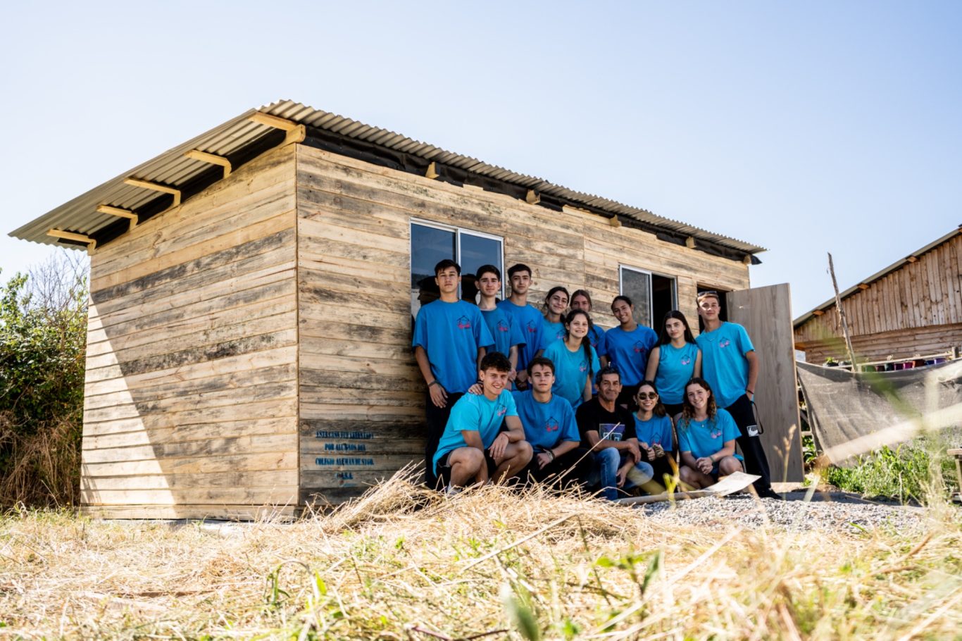Gruppe in blauen T-Shirts posiert vor einem Holzhaus in einer ländlichen Umgebung.