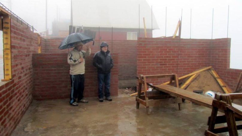 Zwei Personen stehen unter einem Regenschirm auf einer Baustelle mit Ziegelwänden.