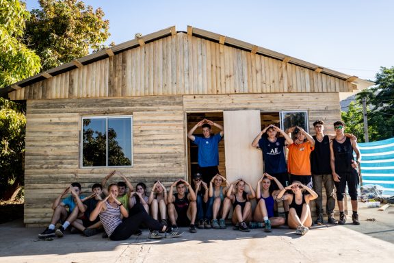 Gruppenfoto vor einem neu erbauten Holzhaus mit jugendlichen Bauarbeitern und Helfern.