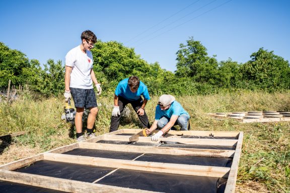 Drei Personen arbeiten im Freien an einem Holzrahmen in einer grünen Landschaft.